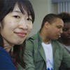 A female student smiling at the camera during a class, with other students interacting in the background.