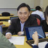 A male student discussing with students during a class, with a laptop and documents on the table.