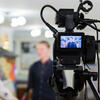 A video camera filming an interview on campus, with a person speaking in the background