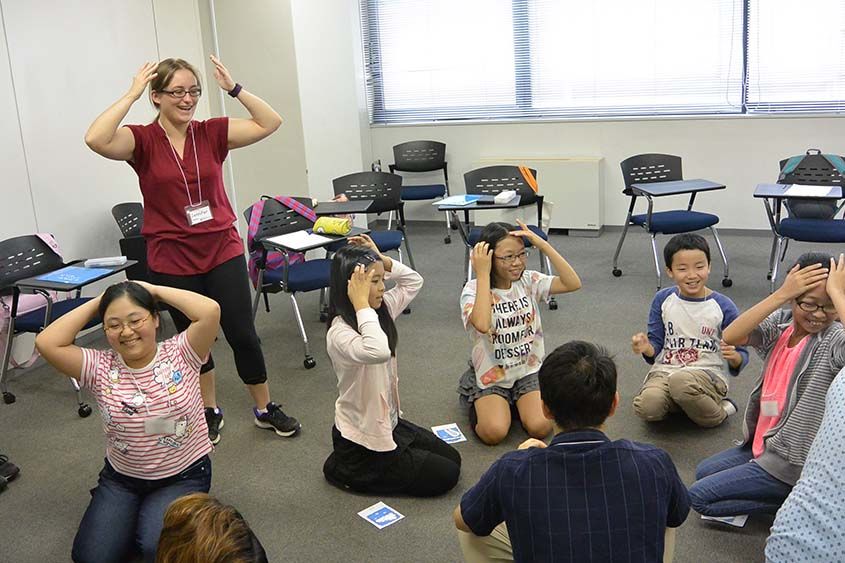 A teacher stands while a group of elementary school students sit in a circle on the floor playing a game